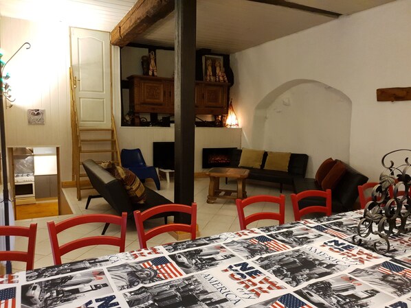 A cozy living room with a fireplace, a dining table with a patriotic tablecloth, and a kitchen area with a stove and cabinets. - ALSACE ,RIQUEWIHR House of the 16th century ( 4 to 14 persons ) (Haut-Rhin)