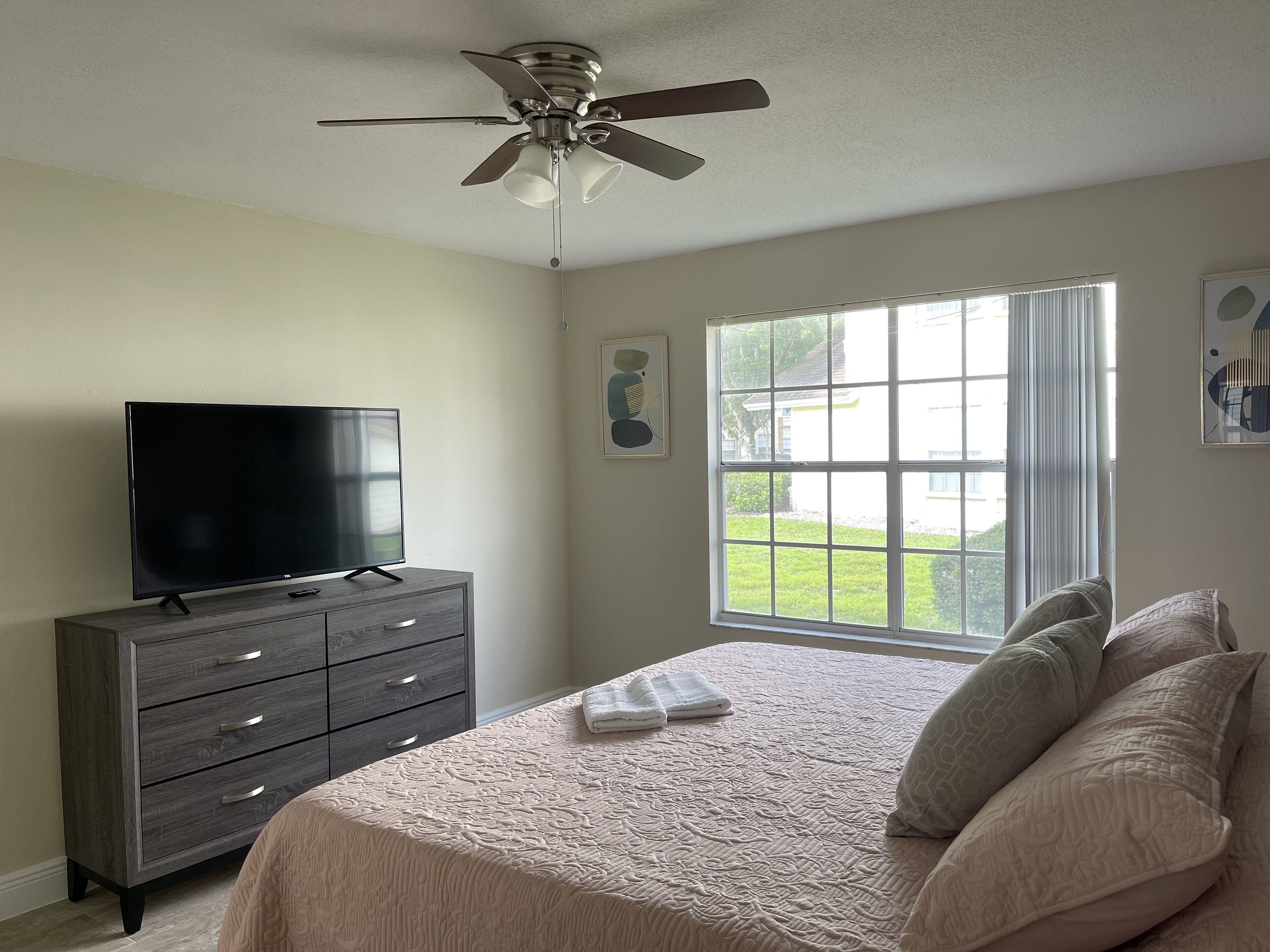 A bedroom with a bed, a TV on a dresser, a ceiling fan, and a window with a view of the outdoors.