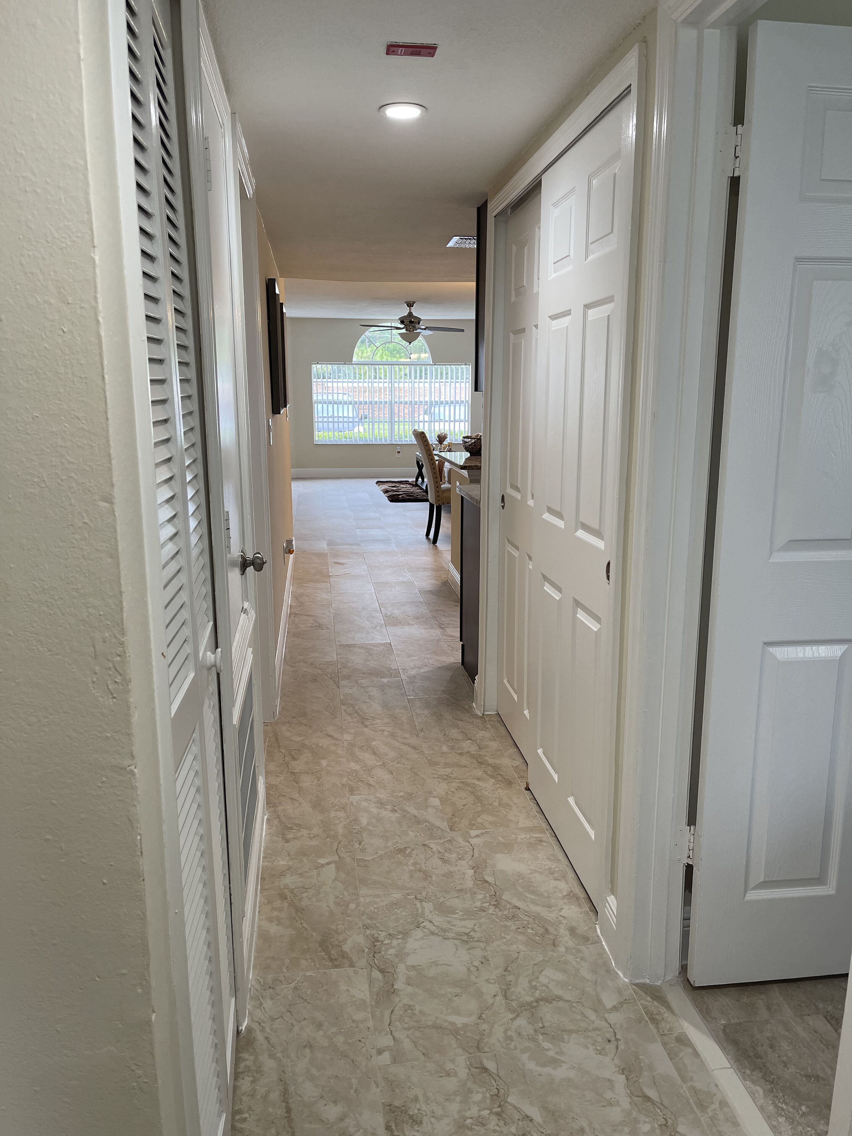 A hallway with a ceiling fan, a dining area with a table and chairs, and a view of a fenced yard through a window.