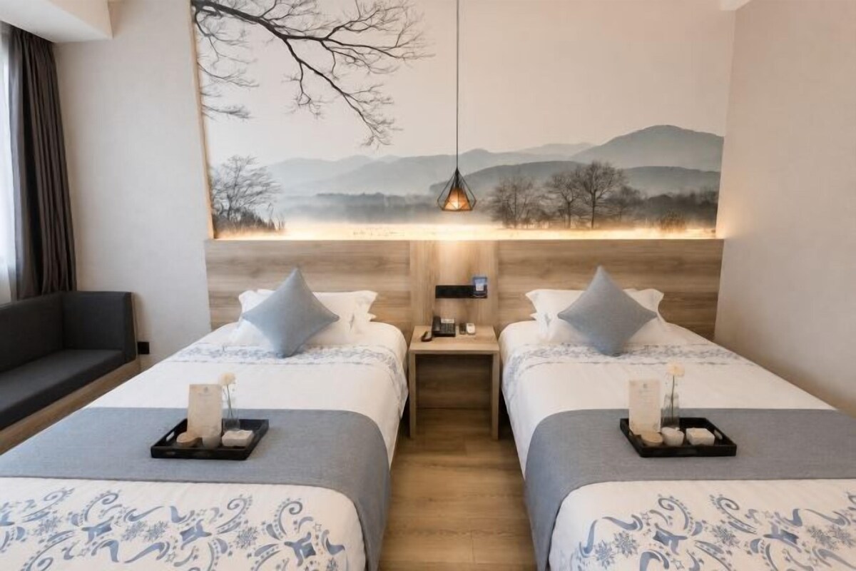 Habitación de hotel con dos camas, una mesita de noche, una lámpara y un cuadro de un paisaje.