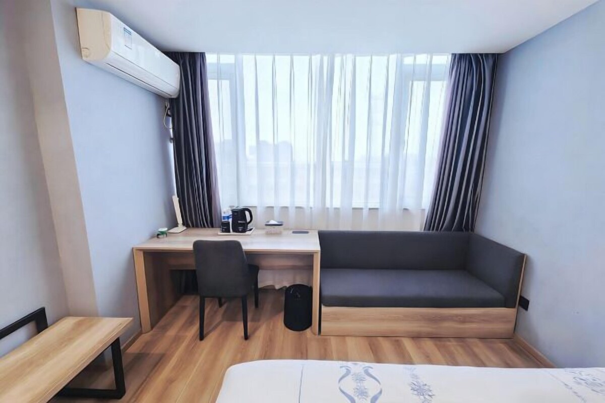 A compact hotel room with a bed, a desk, a chair, a sofa, and a window with curtains.