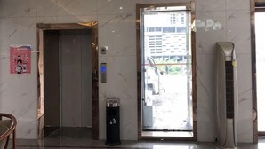 Ascensore, cestino e termosifone in un atrio con pavimento piastrellato e una porta che dà sull'esterno.