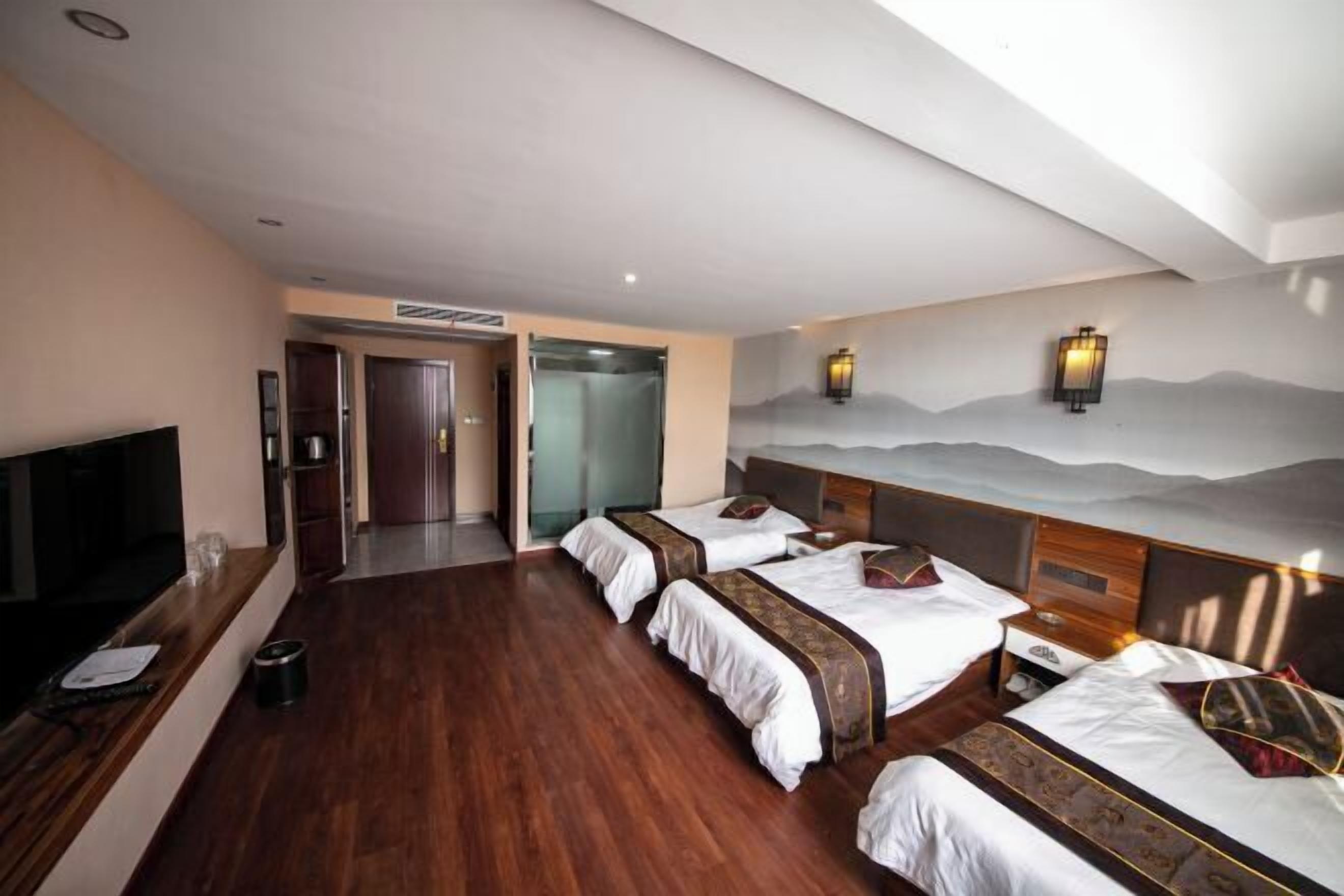 A hotel room with three beds, a TV, a desk, and a wall-mounted lamp.