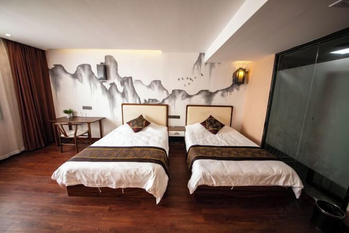 A hotel room with two beds, a wooden floor, a desk, a chair, and a wall with a mountain painting.