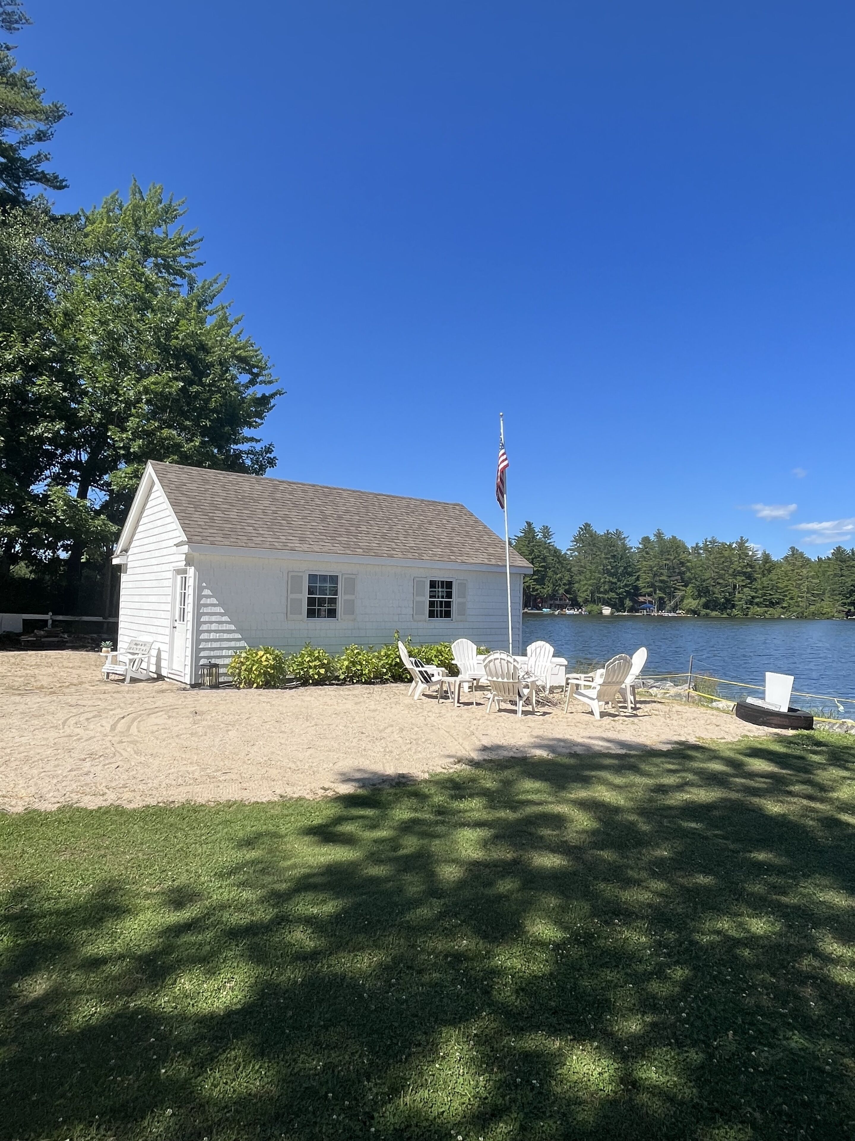 Une maison blanche avec un mât pour drapeau, une jetée et des chaises au bord d’un lac.