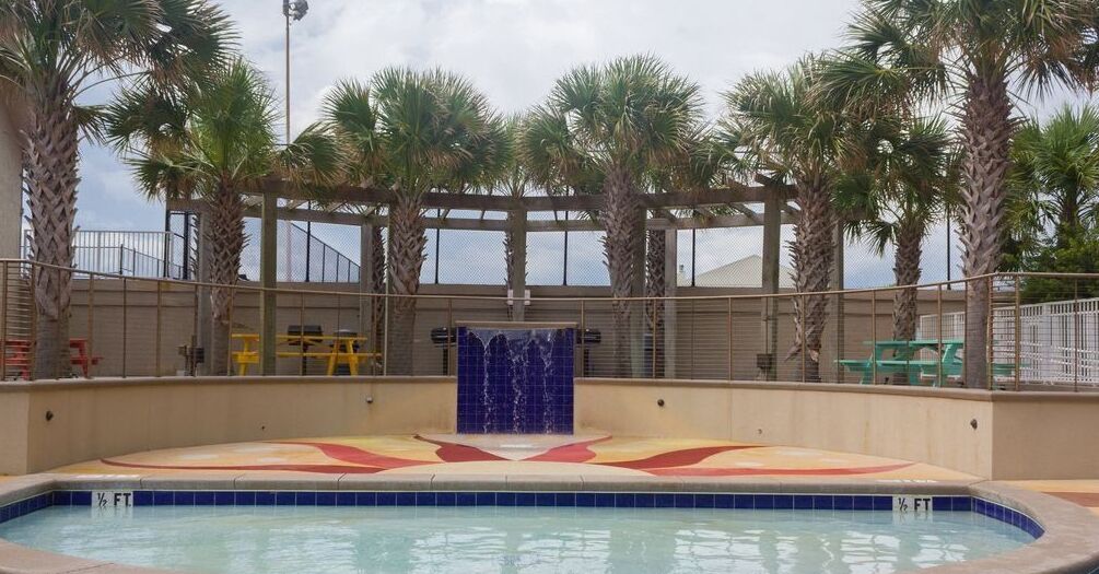 A swimming pool with a blue water feature, surrounded by palm trees and a covered area with a yellow bench.