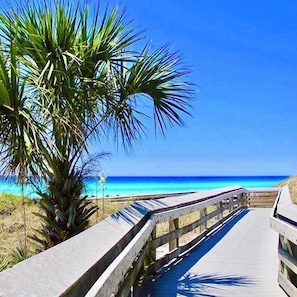Un paseo marítimo de madera que lleva a una playa con agua azul cristalina y una palmera.