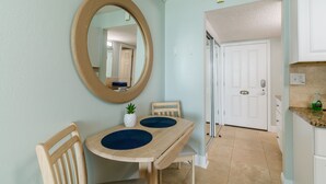 A dining area with a wooden table and chairs, a round mirror on the wall, and a kitchen area with cabinets and a countertop. - Treasure Island Oceanfront Studio with Balcony & Heated Pool (Treasure Island)