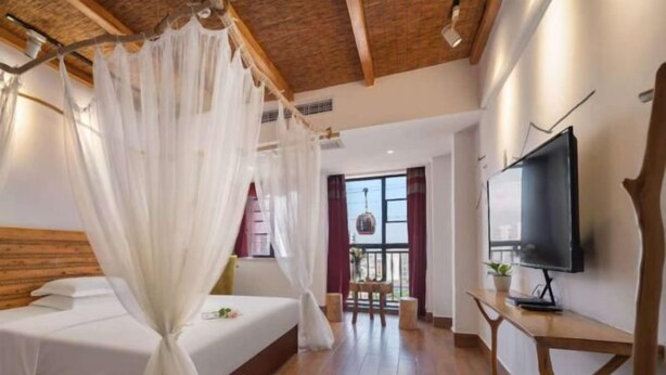 A bedroom with a bed, a TV, a wooden floor, a ceiling with exposed beams, and a window with curtains.