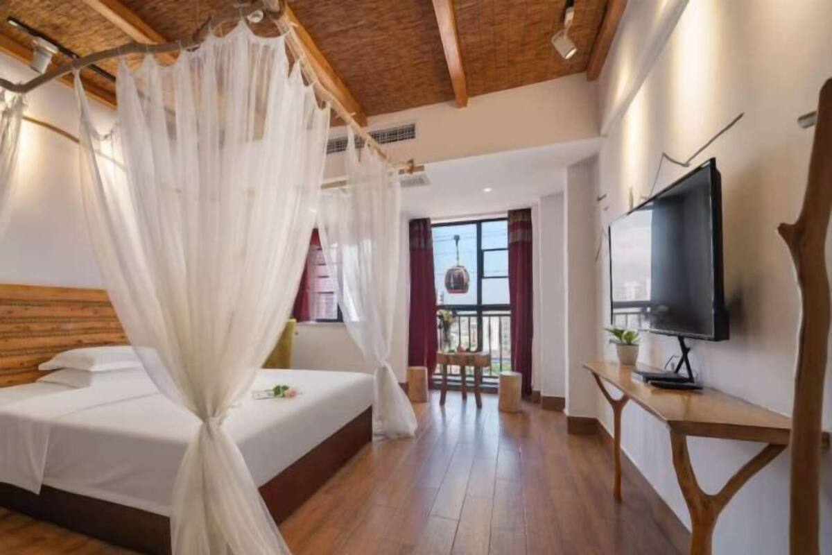 A bedroom with a bed, a TV, a wooden floor, a ceiling with exposed beams, and a window with curtains.