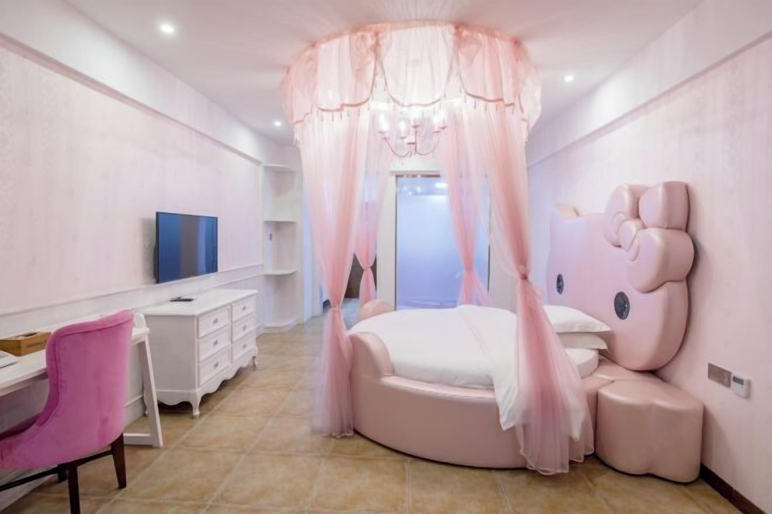 Une chambre à coucher avec un lit rond, une coiffeuse avec un miroir, un fauteuil rose et un téléviseur posé sur une commode blanche.