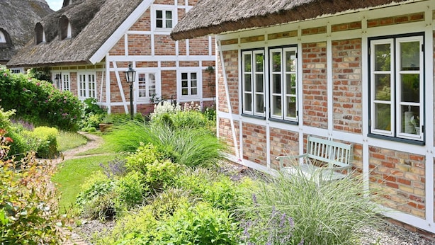 A traditional thatched-roof cottage with a brick exterior, white window frames, and a well-maintained garden with various plants and a pathway. - Landhaus Vilmnitz - Wohnung 9 - Landhaus Vilmnitz, Wohnung 9 (Putbus)
