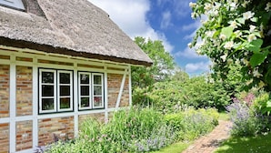 A traditional thatched-roof cottage with a brick exterior and white-framed windows, surrounded by a lush garden with a variety of flowering plants and a brick pathway. - Landhaus Vilmnitz - Wohnung 9 - Landhaus Vilmnitz, Wohnung 9 (Putbus)