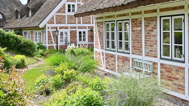 Een traditionele rieten cottage met een bakstenen gevel, witte raamkozijnen en een verzorgde tuin met verschillende planten en een pad.