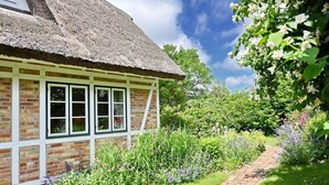 Een pittoresk rieten dak huisje met een bakstenen gevel en ramen met witte kozijnen, omringd door een weelderige tuin vol bloeiende planten en een pad van baksteen.