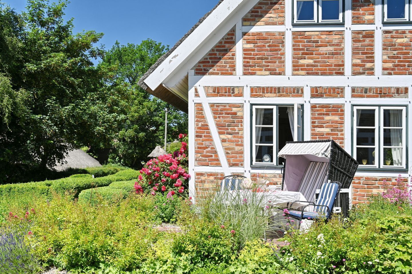 Ein traditionelles Backsteinhäuschen mit weißer Fassade, ein Garten mit verschiedenen Pflanzen und Blumen sowie eine Terrasse mit Stuhl und Tisch.