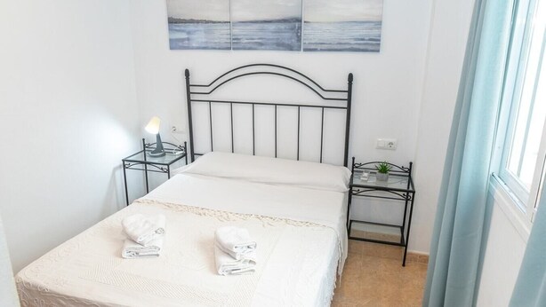 Un dormitorio ordenado con una cama, dos mesitas de noche, una lámpara y un cuadro sobre la cama.