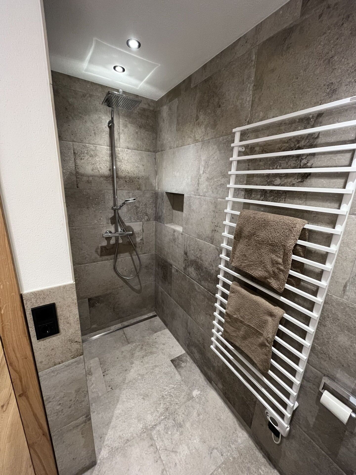 A modern shower with a handheld showerhead, a white towel rack with towels, and a recessed ceiling light.