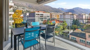 Un balcon avec une table et des chaises, donnant sur une ville de montagne avec ses bâtiments et sa végétation.