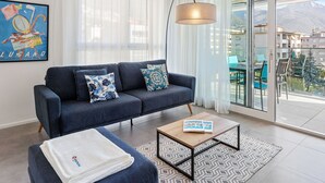 Un salon moderne avec un canapé bleu foncé, des coussins à motifs, une table basse et une grande fenêtre donnant sur des immeubles et des montagnes.