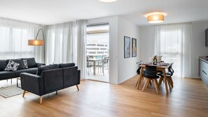Un saló modern amb un sofà, taula de menjador i caires, una zona de cuina i finestres grans amb cortines.