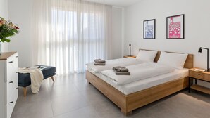 A modern hotel room with a large bed, a bedside table with a lamp, a small desk with a chair, a dresser, and a large window with sheer curtains. - Lovely apartment for 2 people with WIFI, TV, balcony and pets allowed (Locarno)