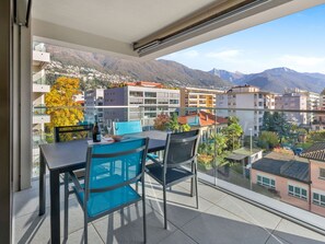 Un balcón con mesa y sillas, con vista a un pueblo montañoso con edificios y vegetación.