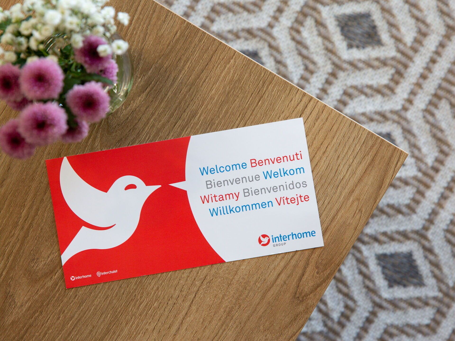 A business card with multilingual welcome message and logo on a wooden surface.