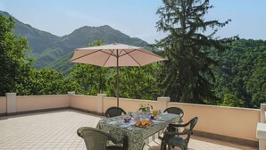 Terraço com mesa posta para refeições, rodeado de cadeiras e um guarda-sol para dar sombra.