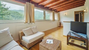 Um quarto amplo com teto de madeira, uma janela grande com cortinas, um sofá, uma televisão sobre um suporte de madeira e uma mesa pequena.