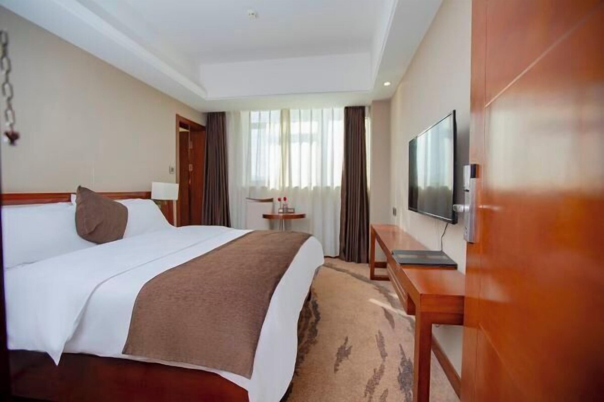 A hotel room with a large bed, a TV, a desk, and a window with curtains.