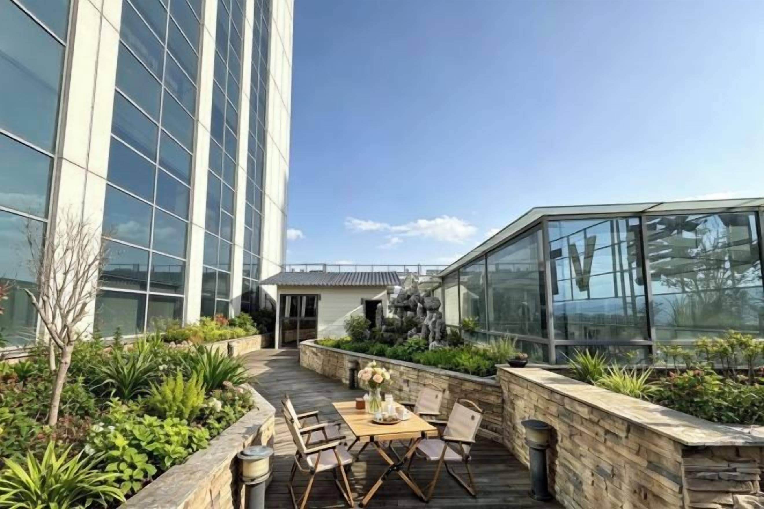 Terraza en la azotea con mesa y sillas, rodeada de vegetación y un edificio de cristal.