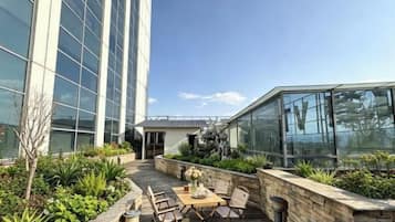 Terraza en la azotea con mesa y sillas, rodeada de vegetación y un edificio de cristal.