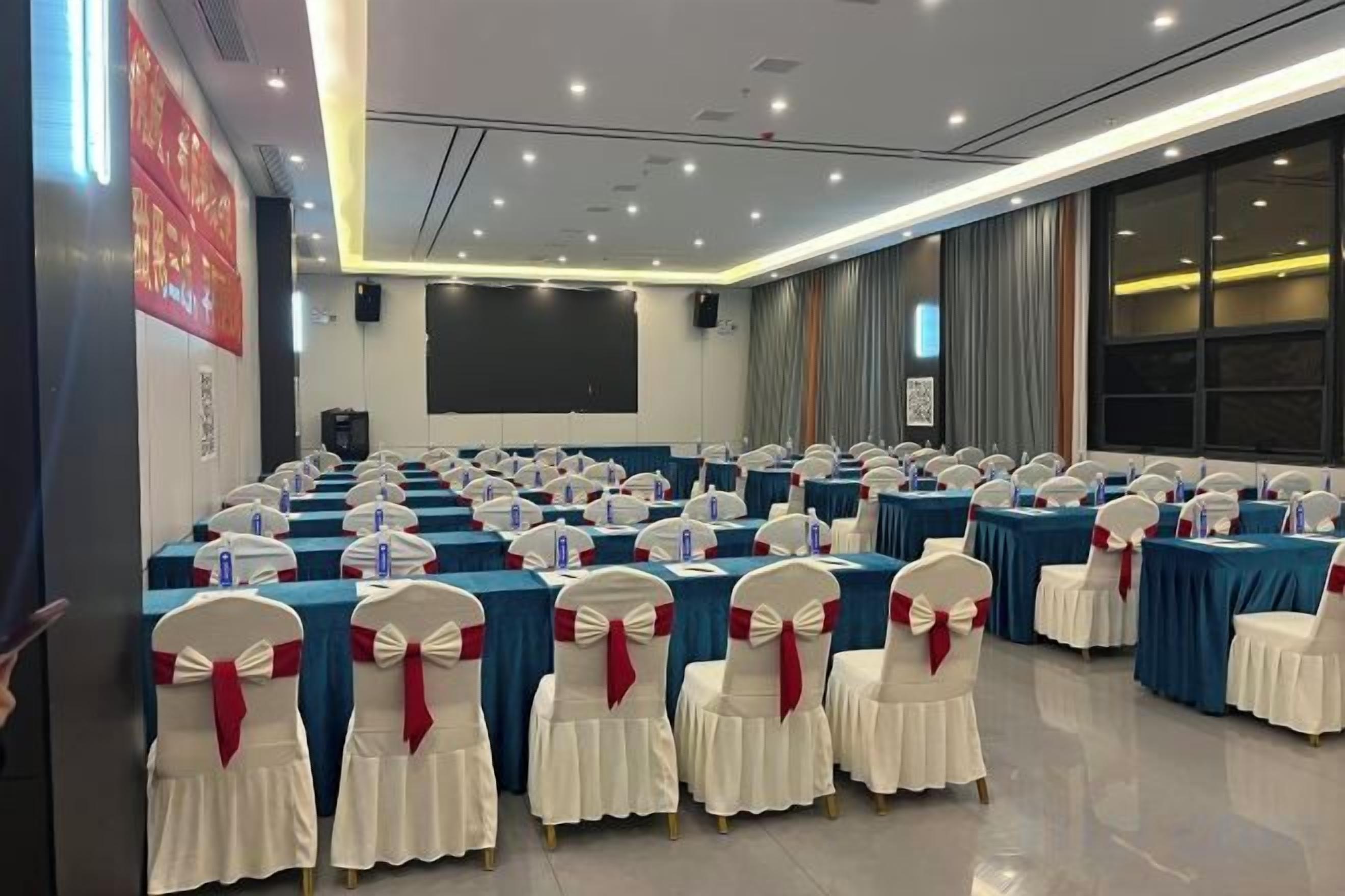 Une salle de conférence avec des rangées de tables et de chaises, un écran de projection et une grande fenêtre.