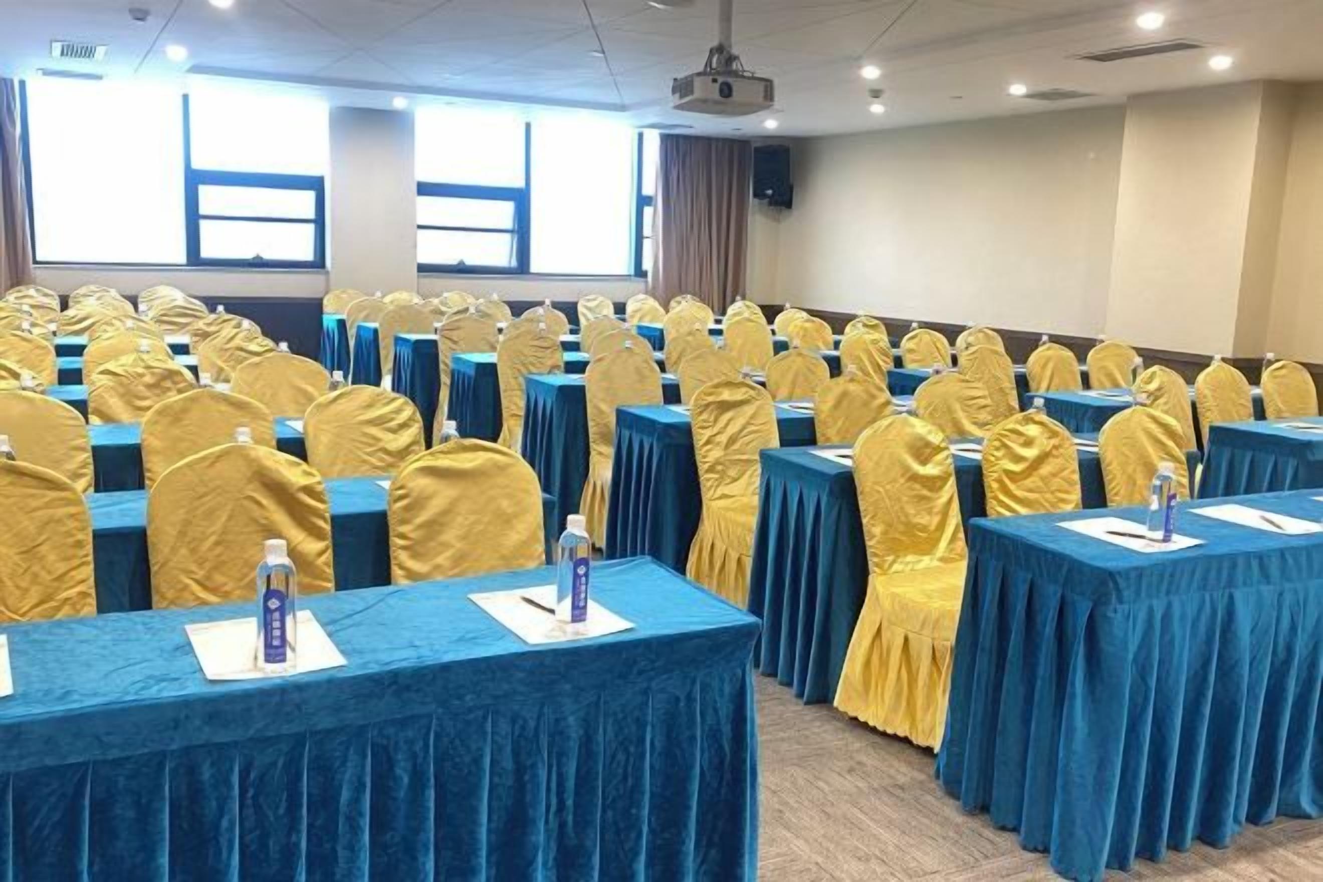 Un salón de conferencias con filas de mesas cubiertas con manteles azules y fundas amarillas para las sillas, equipado con botellas de agua y tarjetas de identificación.