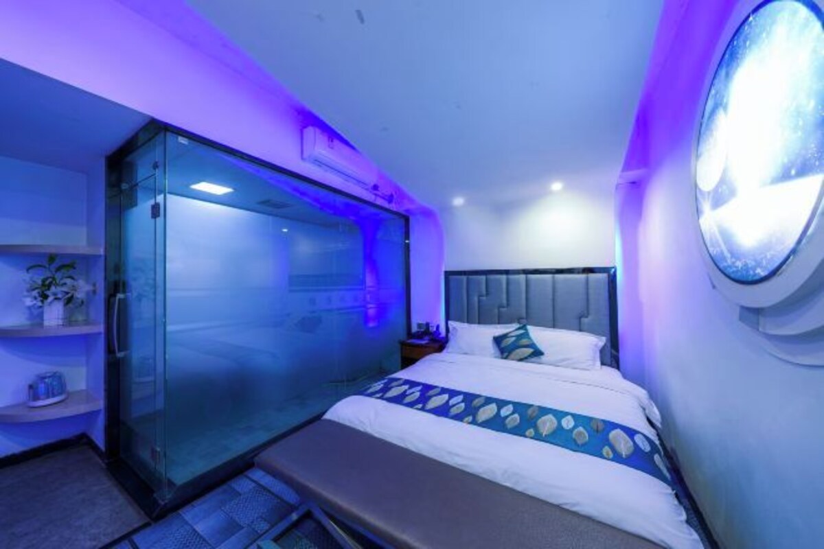 A modern hotel room with a glass-enclosed shower, a bed with a patterned bedspread, and a round mirror on the wall.