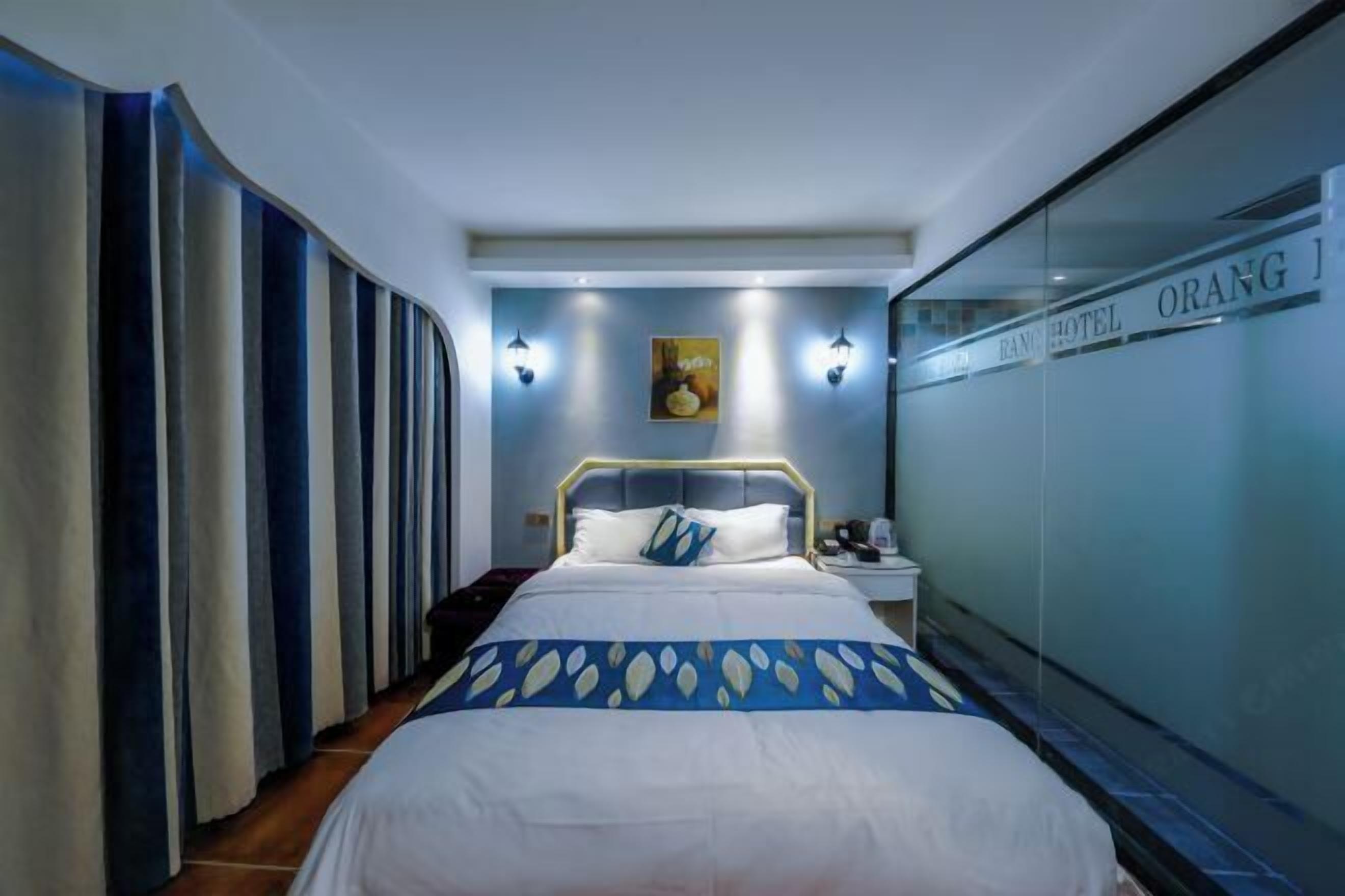 A hotel room with a bed, a nightstand, a wall-mounted lamp, and a glass partition with the hotel name.