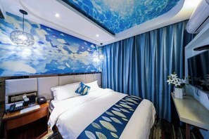 A hotel room with a bed, a nightstand, a desk, a chair, a wall with a blue abstract design, and a curtain.