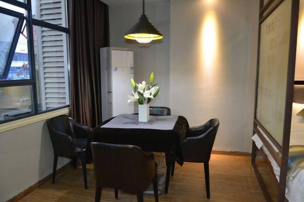 Una habitación con una mesa de comedor, sillas, un jarrón con flores, una lámpara en el techo y una cama con cabecera.
