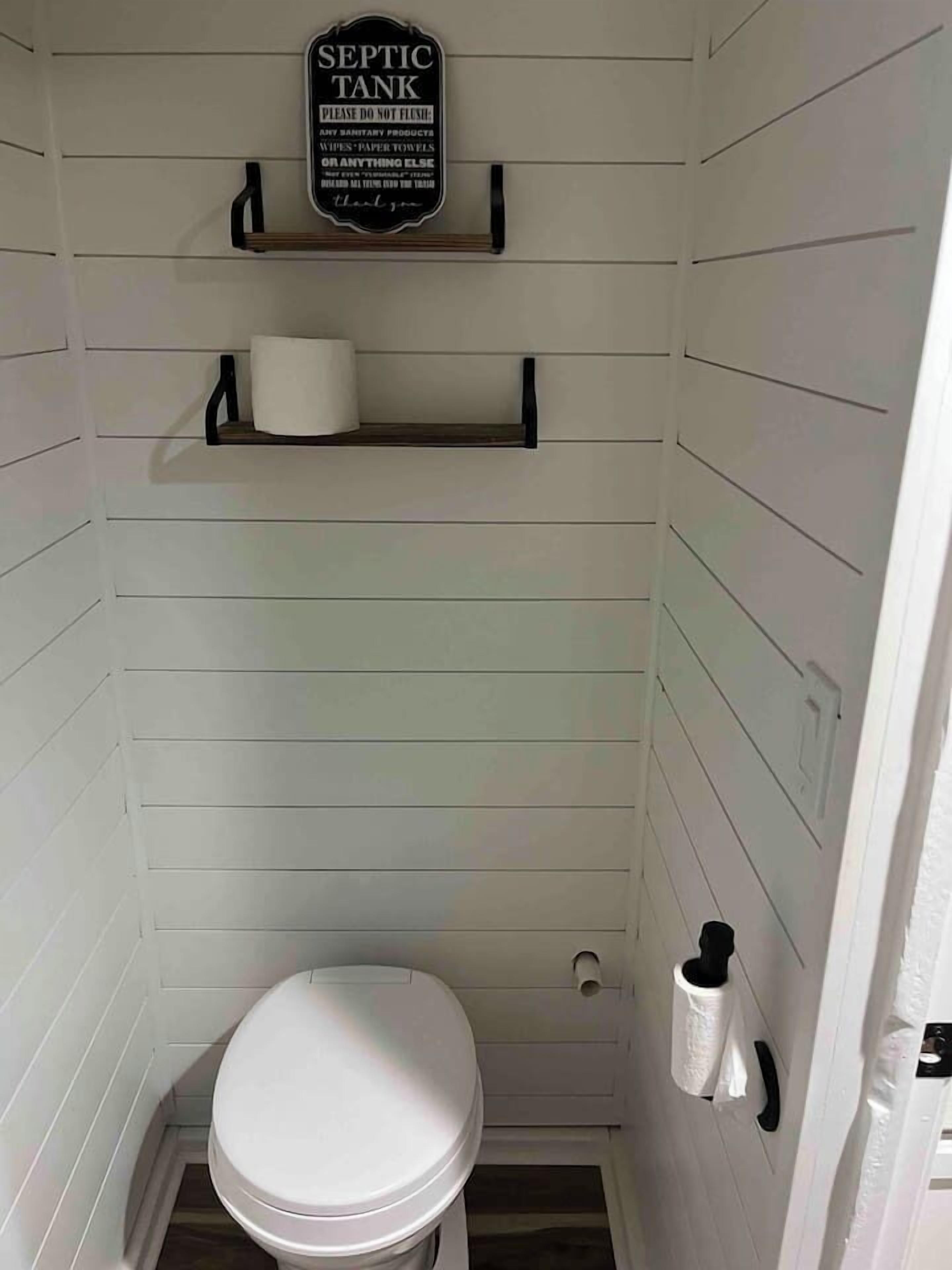 Ein kleines Badezimmer mit weißen Paneelen, ausgestattet mit Toilette, Toilettenpapierhalter und einem Schild über der Toilette, das darauf hinweist, keine Hygieneartikel zu entsorgen.