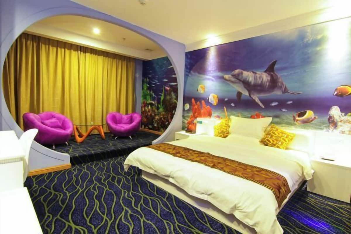 A hotel room with a bed, a large window with yellow curtains, a mural of an underwater scene, and two purple chairs.