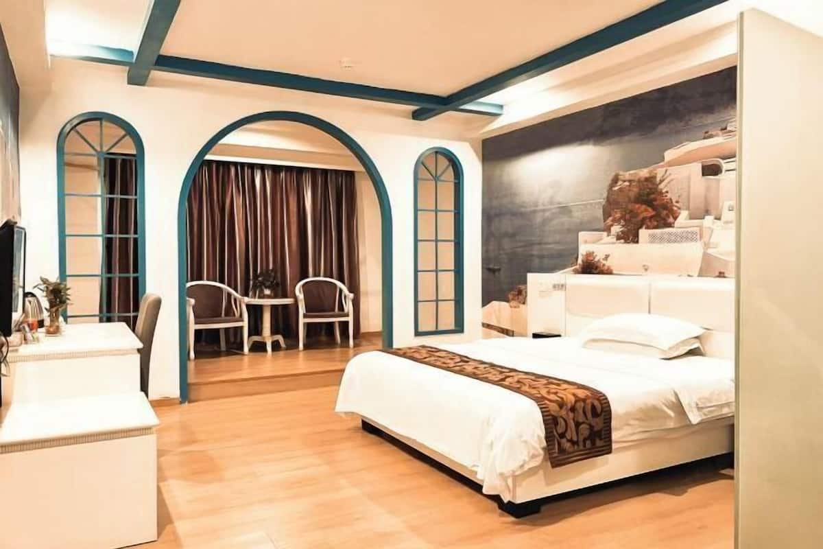 A modern hotel room with a large bed, a desk, and a chair. There are two arched windows and a dark wall with a decorative piece.