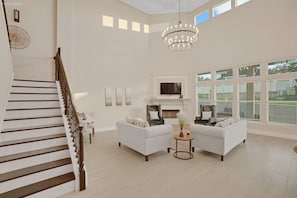 Amplia sala de estar con escalera, dos sofás, chimenea y ventanas grandes.