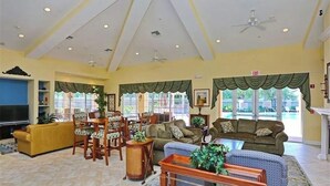 Un salon spacieux avec un canapé jaune, un canapé bleu à motifs et une table basse en bois. Il y a deux fenêtres avec des rideaux verts, un ventilateur de plafond et un téléviseur sur le côté gauche.