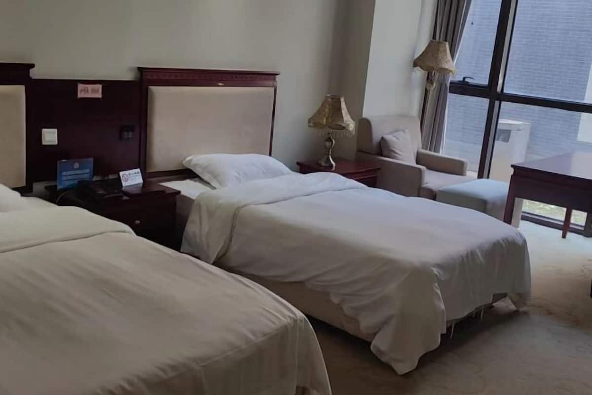 A hotel room with two beds, a desk, a chair, a lamp, and a view of the city through large windows.