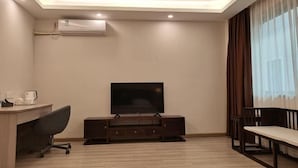 A hotel room with a flat-screen TV on a wooden stand, a desk with a chair, a dining table with a bench, and a large window with curtains. - Hancun River Villa (Beijing)