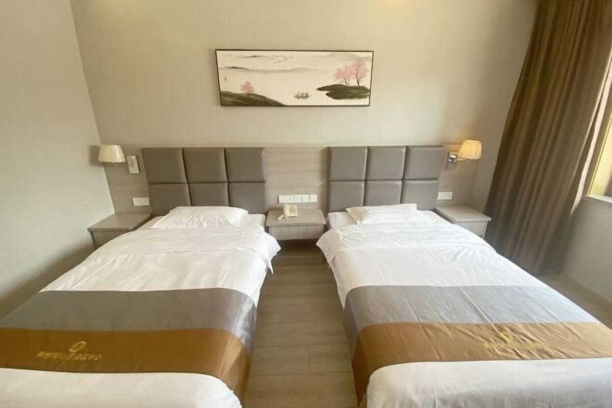 A hotel room with two beds, a headboard, bedside lamps, a painting, and a window with curtains.