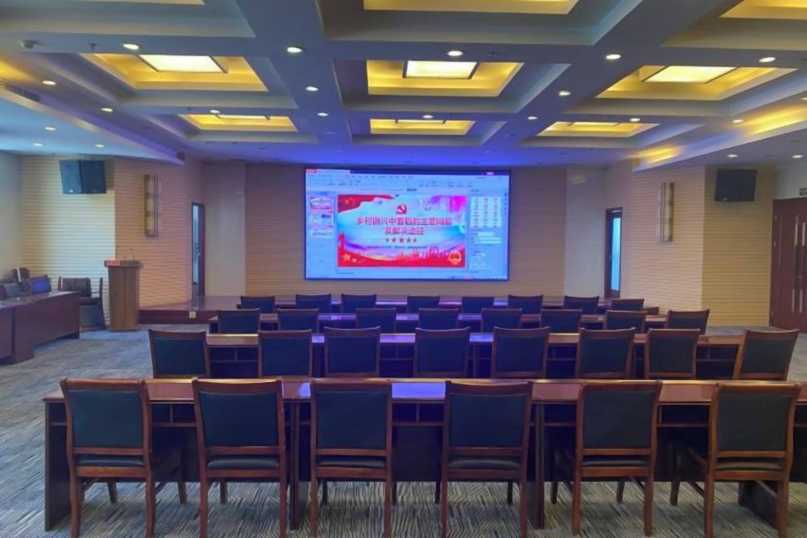 A conference room with rows of chairs facing a large screen displaying a presentation.
