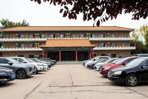 A building with a traditional architectural style, featuring a central entrance with a tiled roof and multiple balconies. There is a parking lot in front with several cars parked. - Hancun River Villa (Beijing)