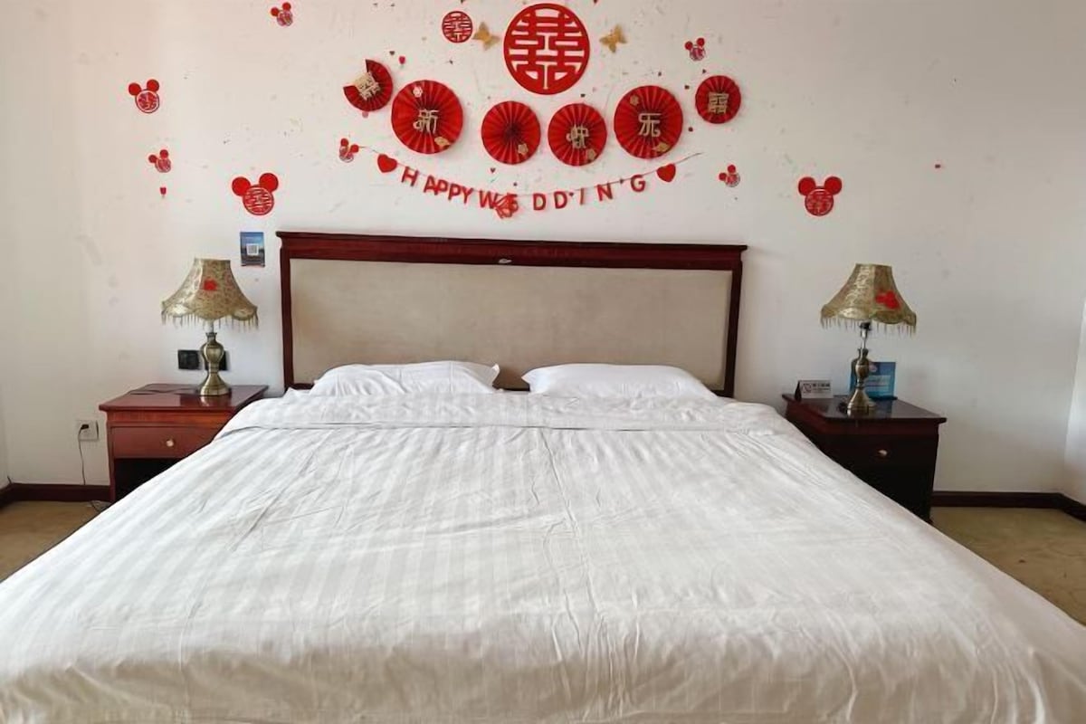 A hotel room with a large bed, two bedside tables with lamps, and a wall decorated with red and gold elements, including a 'Happy Wedding' banner.